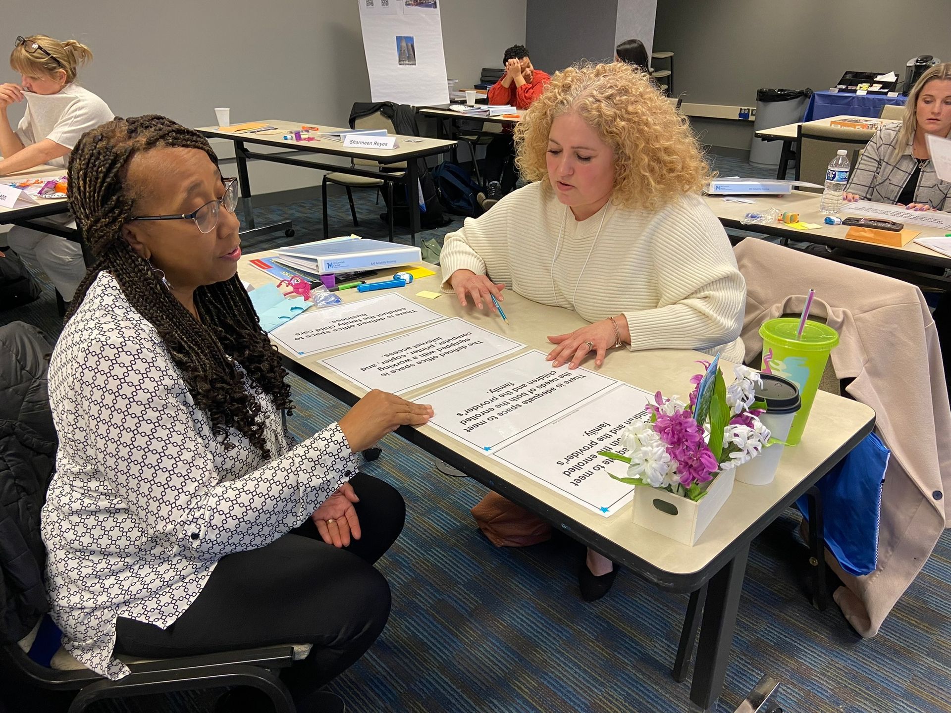 Two women at a table, discussing a document. One points, the other looks on. Indoors, workshop setting.