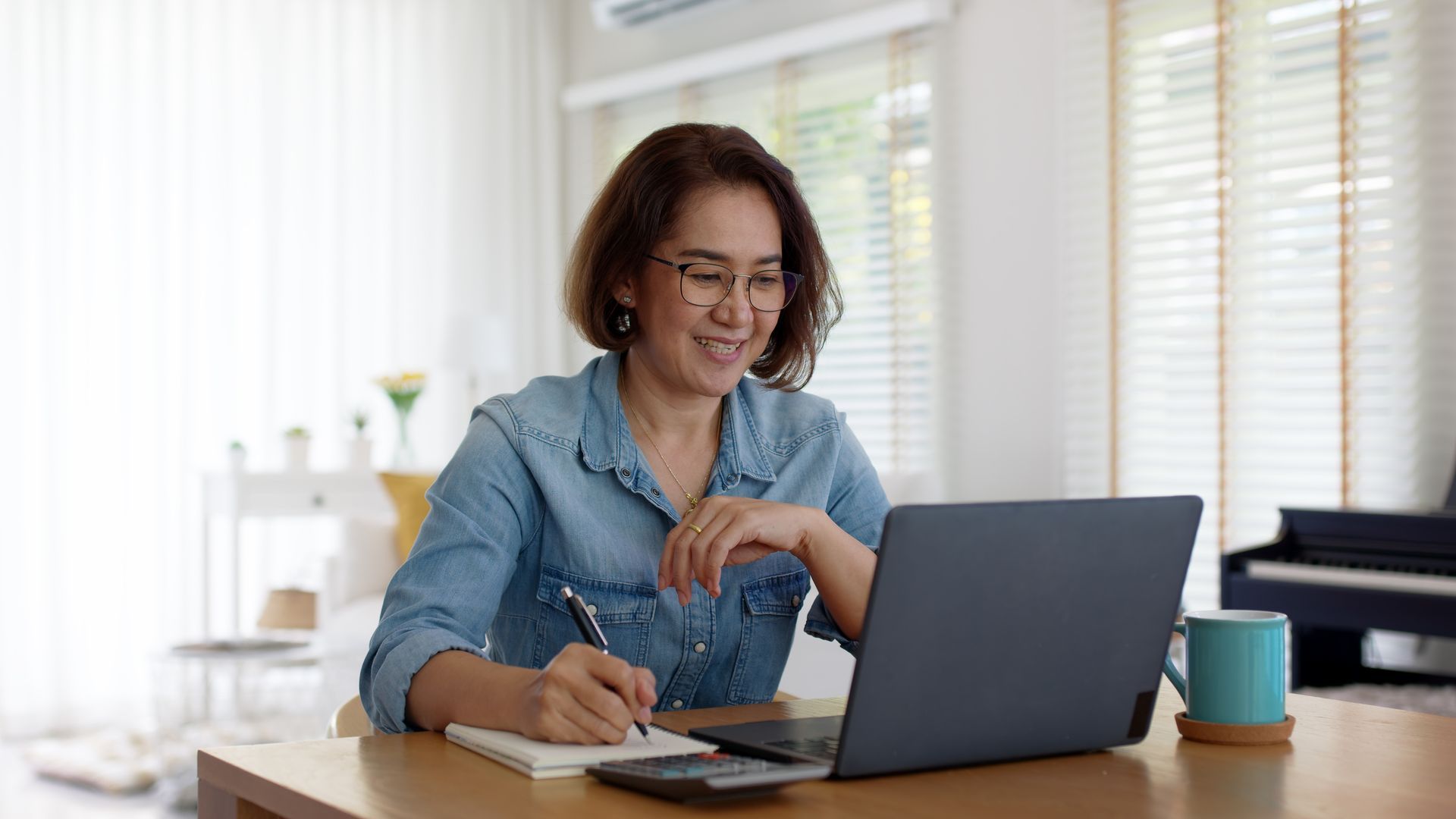 Woman at desk working on laptop, writing in notebook. Smiling, interior setting.