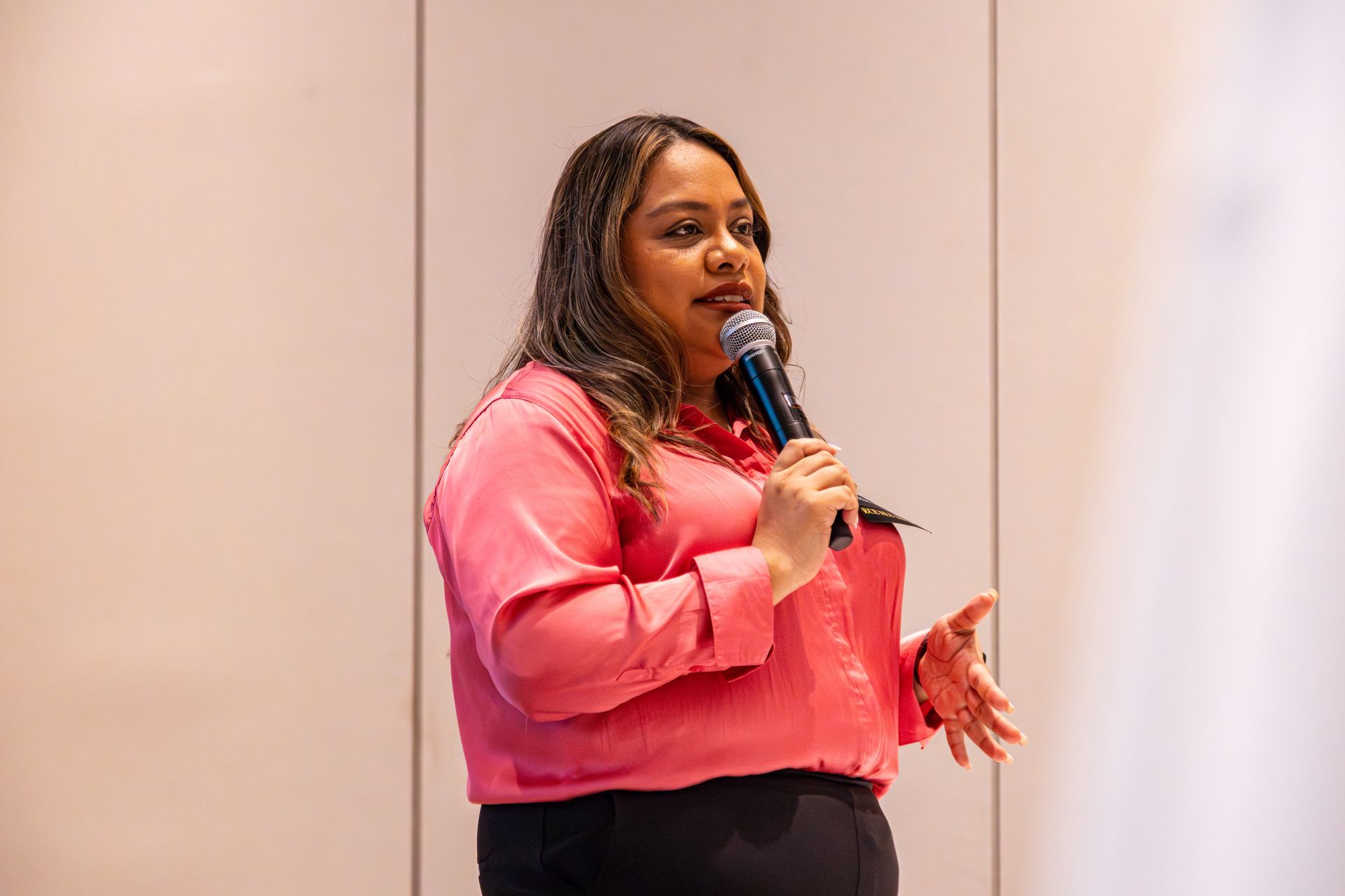 A person in a pink button-down shirt speaking into a microphone while standing in front of a white wall.