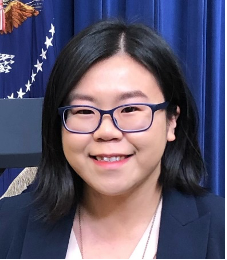 A woman wearing glasses and a suit is smiling in front of a flag.