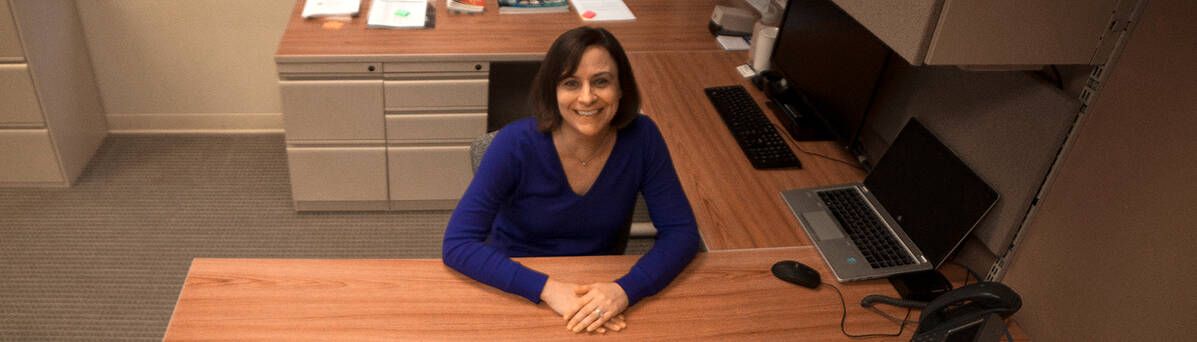 A woman is sitting at a desk in an office with her hands folded.