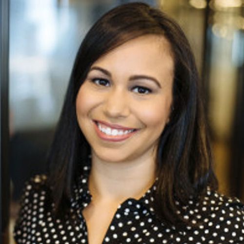 A smiling person with dark hair wears a black and white polka-dot collared shirt in an office setting.