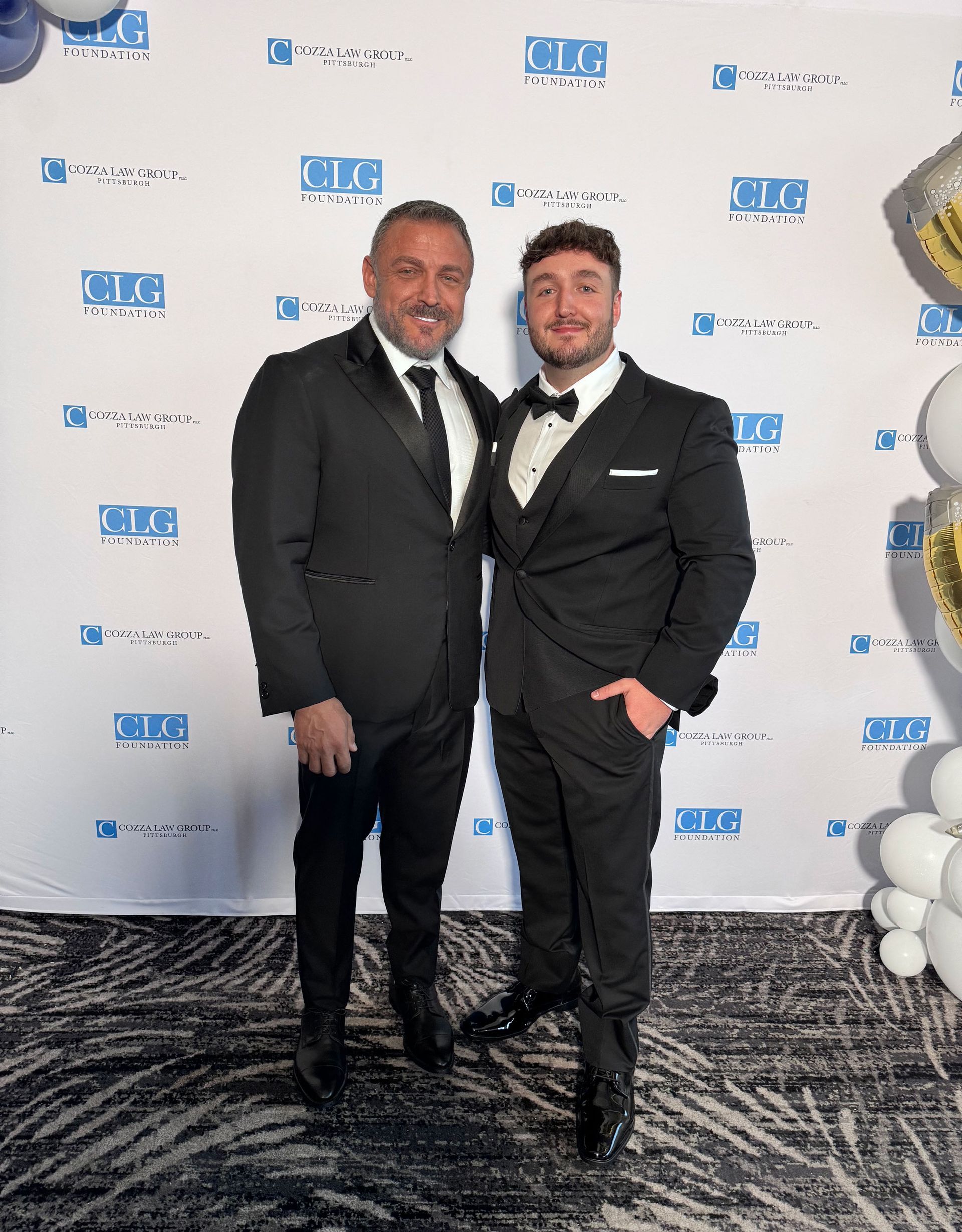 Two men in black suits pose together at an event with a patterned floor and logo backdrop.