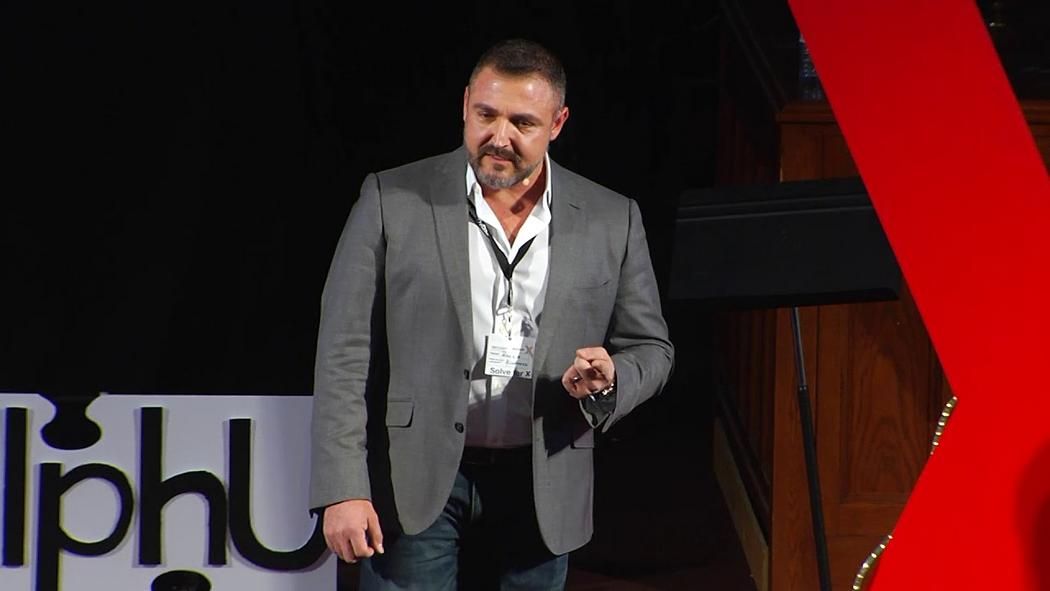 Man in grey blazer on stage, speaking at a TEDx event.