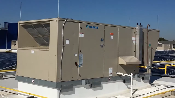 A light tan industrial HVAC rooftop unit installed on a building's flat, light-colored roof against a clear blue sky.
