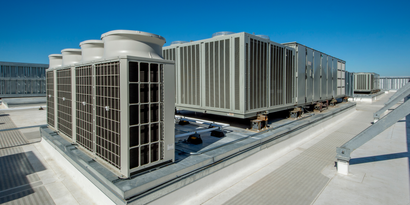 Large commercial HVAC cooling units arranged in rows on a flat building roof under a clear blue sky.