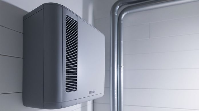 A wall-mounted grey air conditioner or ventilation unit is positioned on a white brick wall near metal pipes.