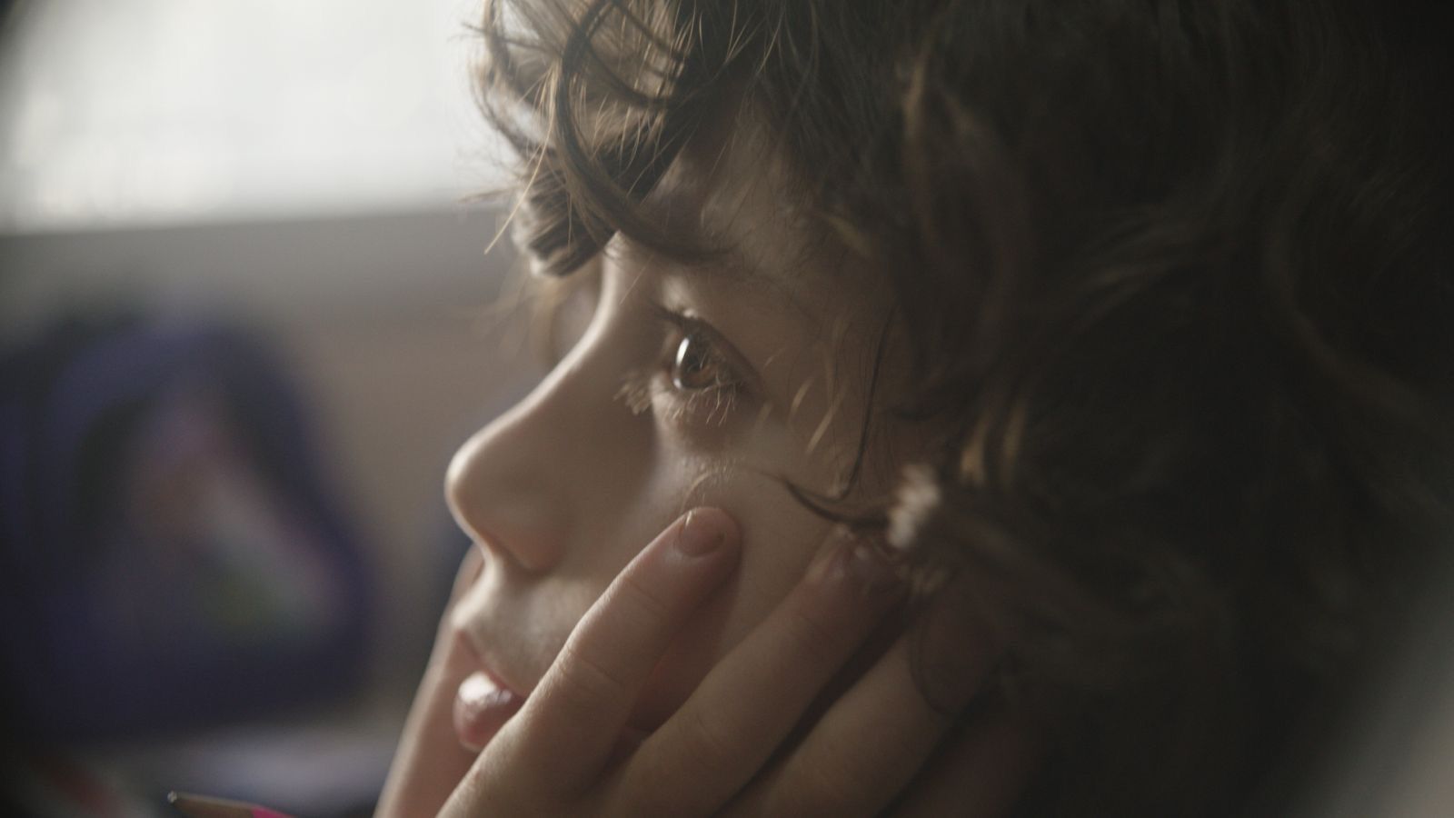 Close-up of a person resting their face on their hand, gazing out a window in soft light.