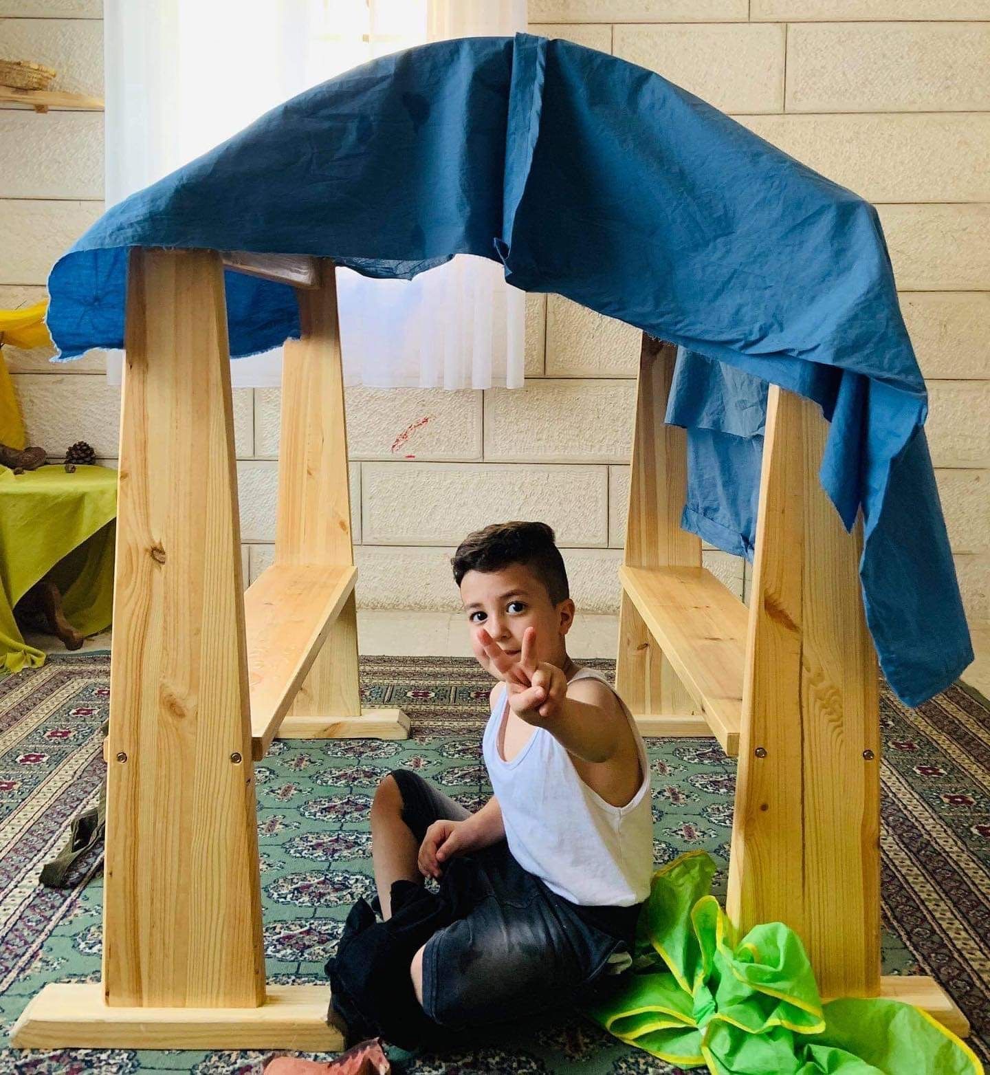 A young boy is sitting under a wooden structure and shows a peace sign with two fingers.