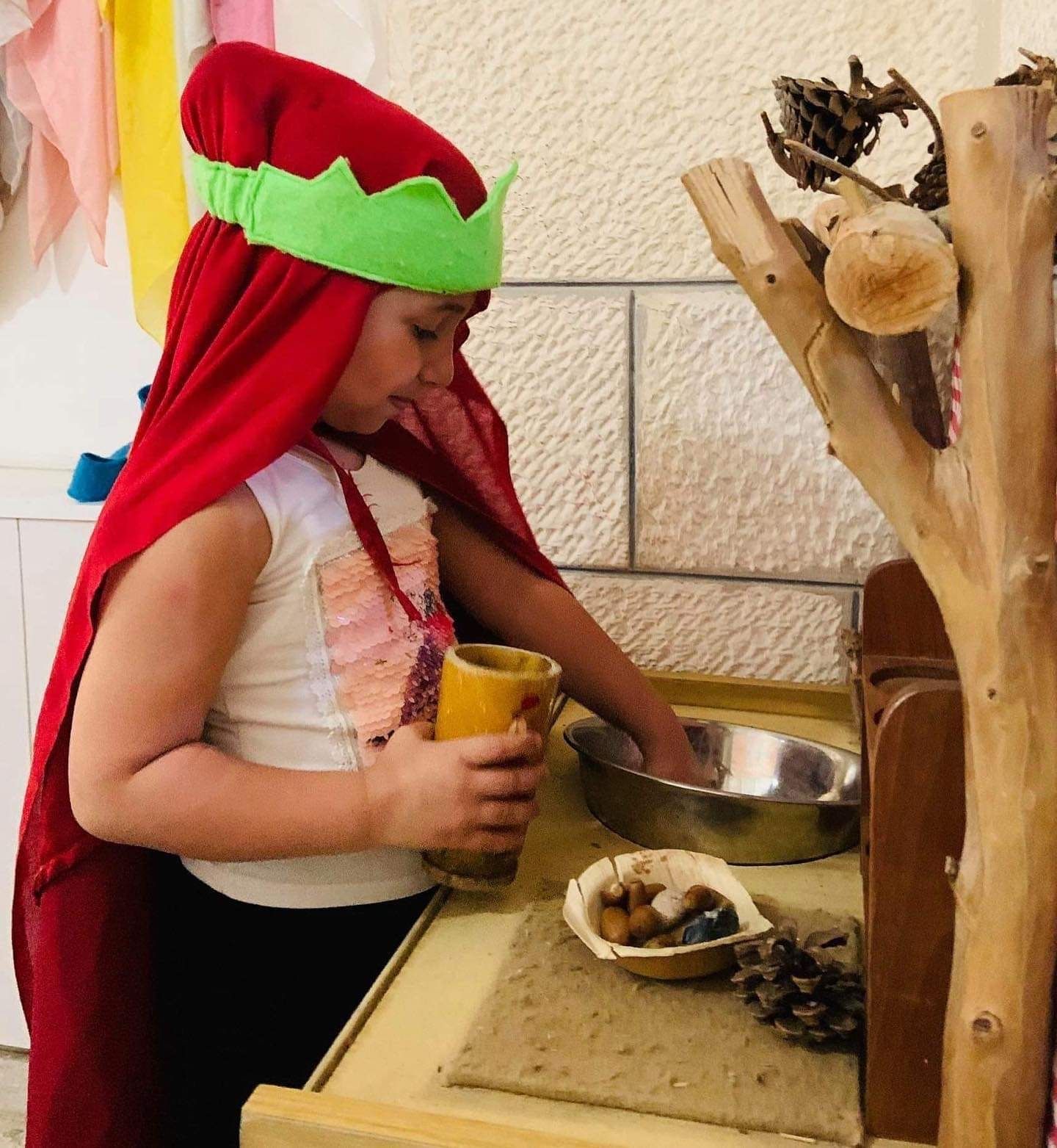 A little girl wearing a red cape and a green crown is sitting at a table holding a cup.