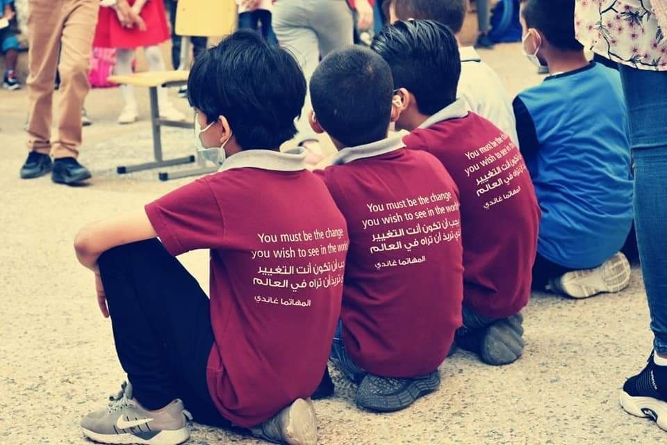 A boy outside, wearing a mask and a maroon shirt with a shield on it, with his elbows out.