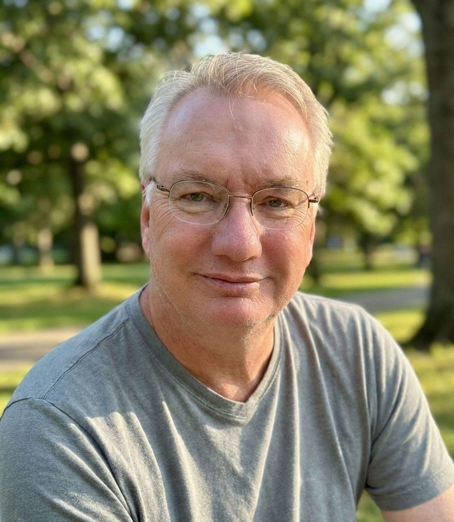 Man with glasses in a gray shirt smiles outdoors in front of a blurred green background.