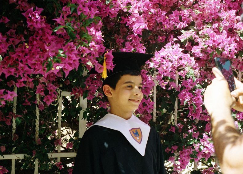 A young boy wearing a graduation cap and gown is standing in front of a bush of pink flowers.