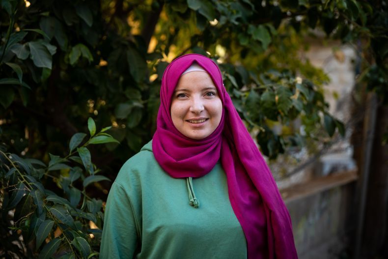 A woman wearing a pink hijab and a green shirt is standing in front of a tree.