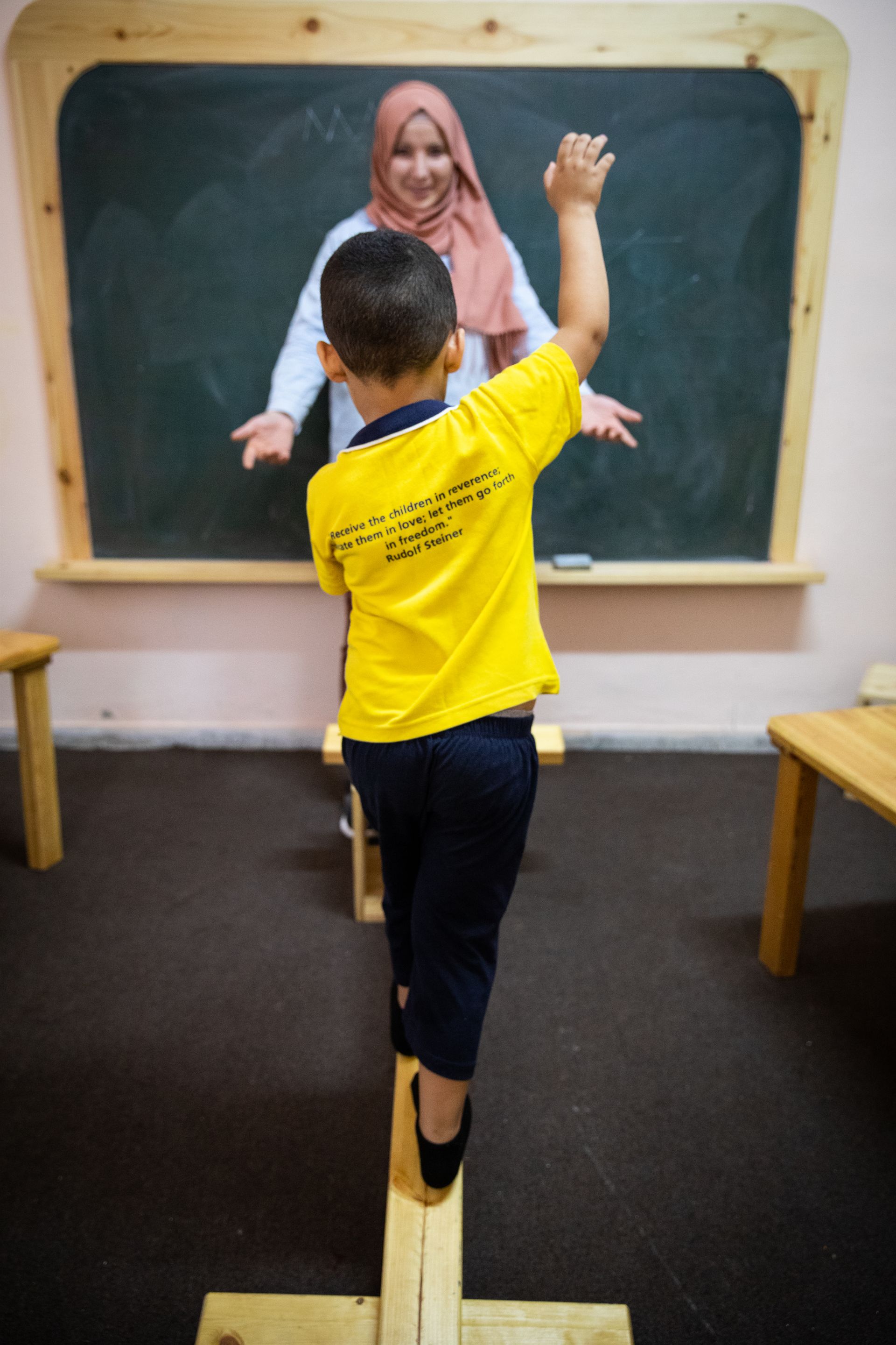 A young boy is balancing on a wooden balance beam in front of a teacher.