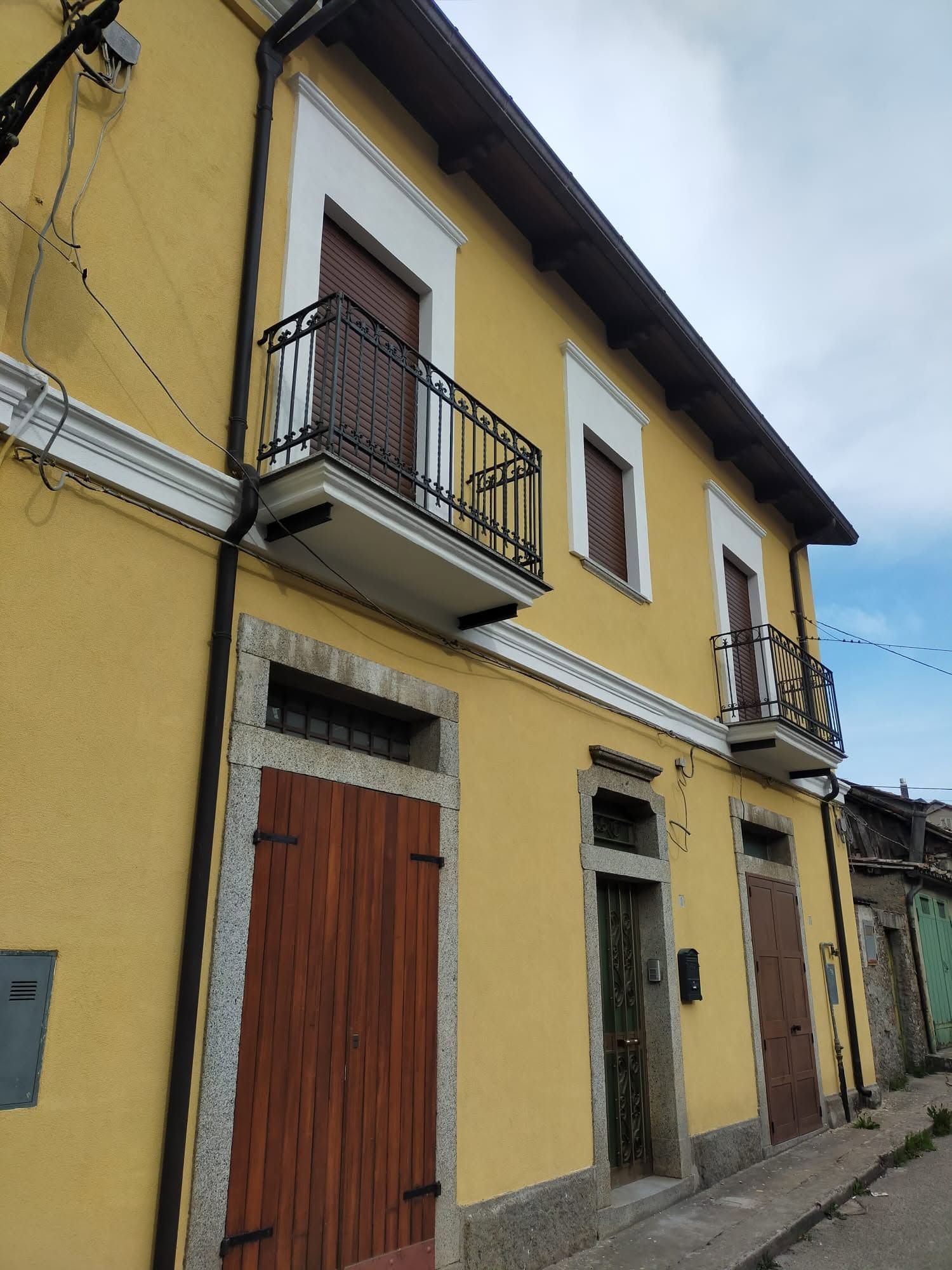 Edificio stradale giallo a due piani con balconi, porte in legno e un marciapiede stretto.