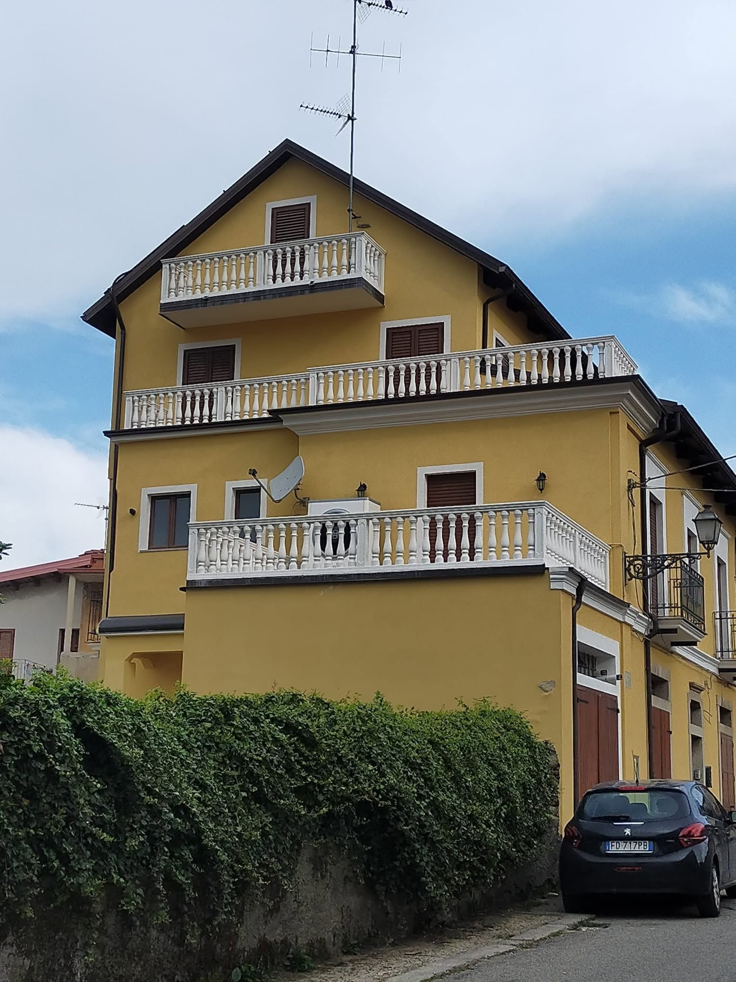 Casa gialla con balconi bianchi e un'auto nera parcheggiata accanto a un muro ricoperto di edera