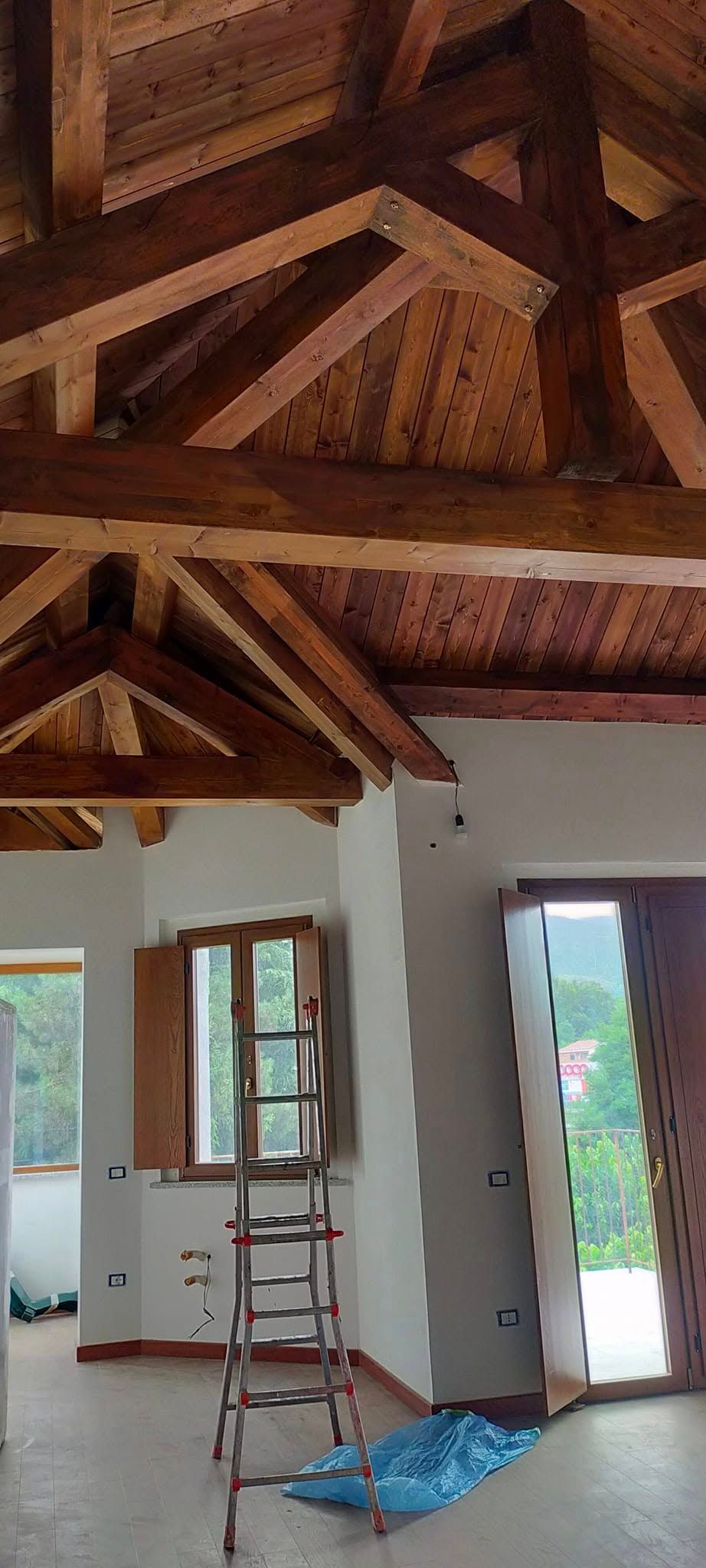 Interno con travi in legno a vista sul soffitto, porta aperta, scala e materiali di ristrutturazione sul pavimento.
