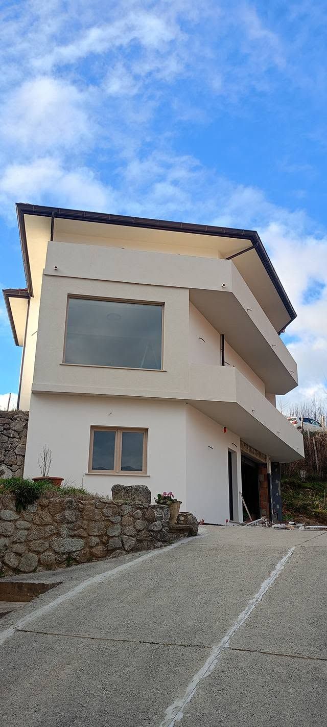 Casa bianca a due piani con grandi finestre, muro di contenimento in pietra e vialetto in pendenza sotto un cielo azzurro