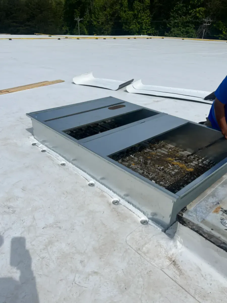 A partially opened rooftop access hatch on a white flat roof. A person is nearby.
