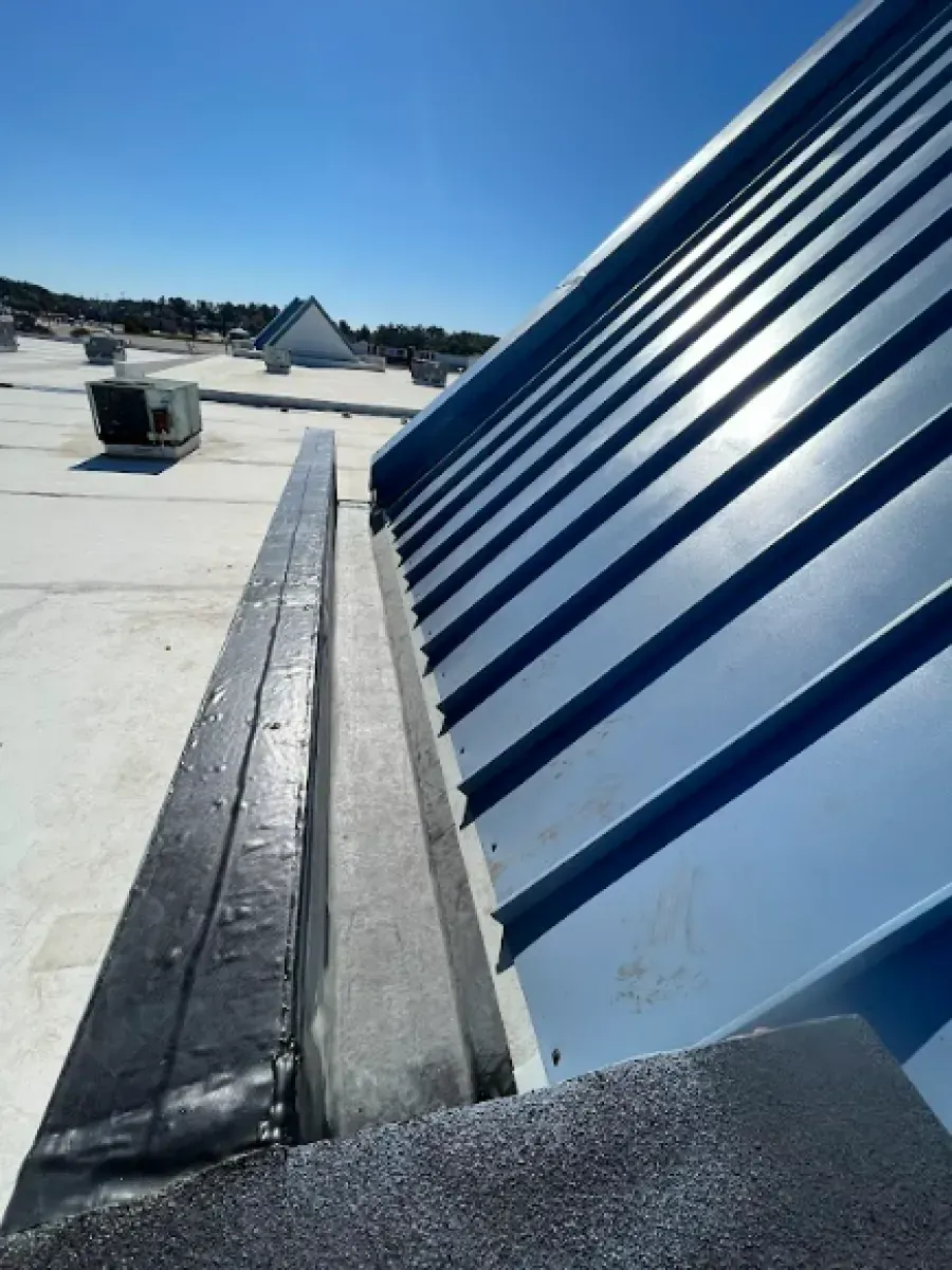 Blue metal roof edge meets a gray gutter, atop a white flat roof under a blue sky.