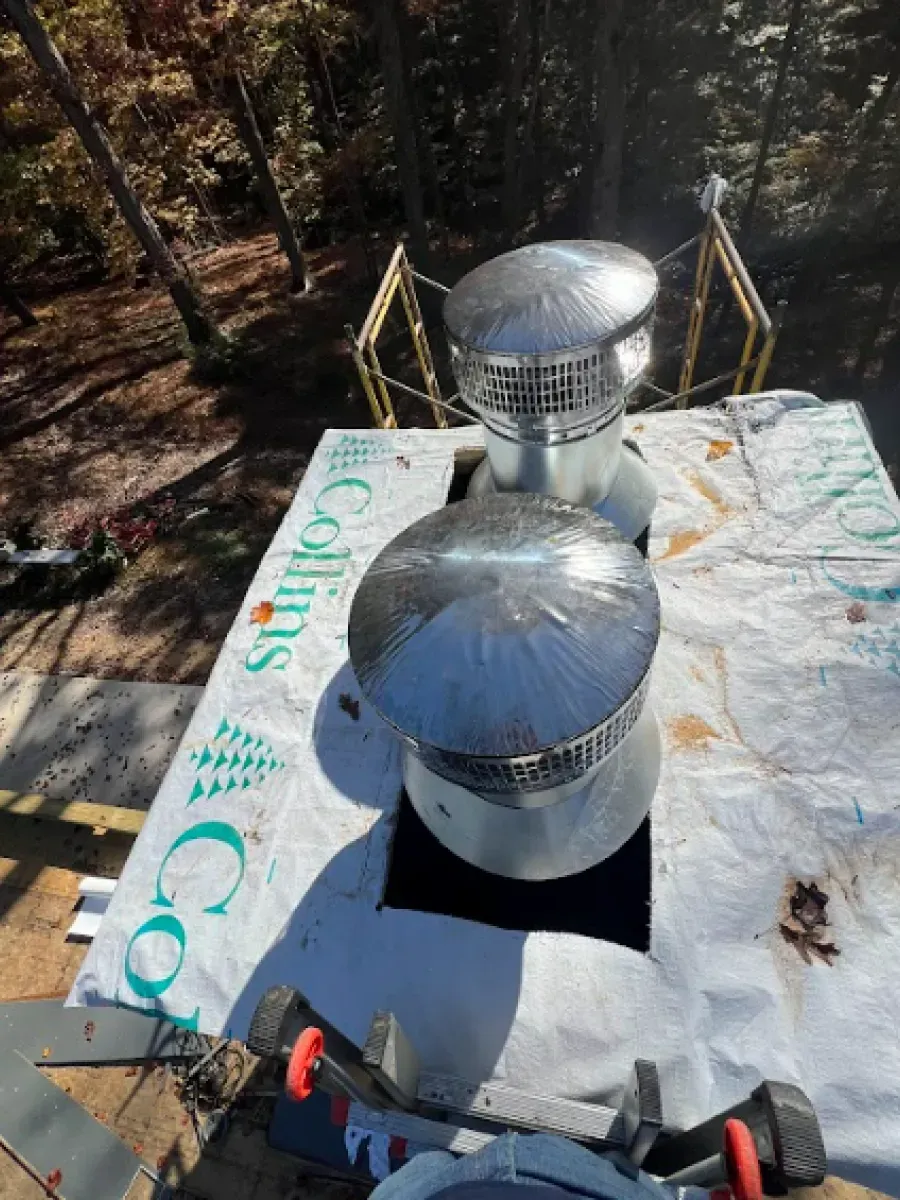 Two silver roof vents on a white surface, surrounded by construction materials, set in a wooded area.