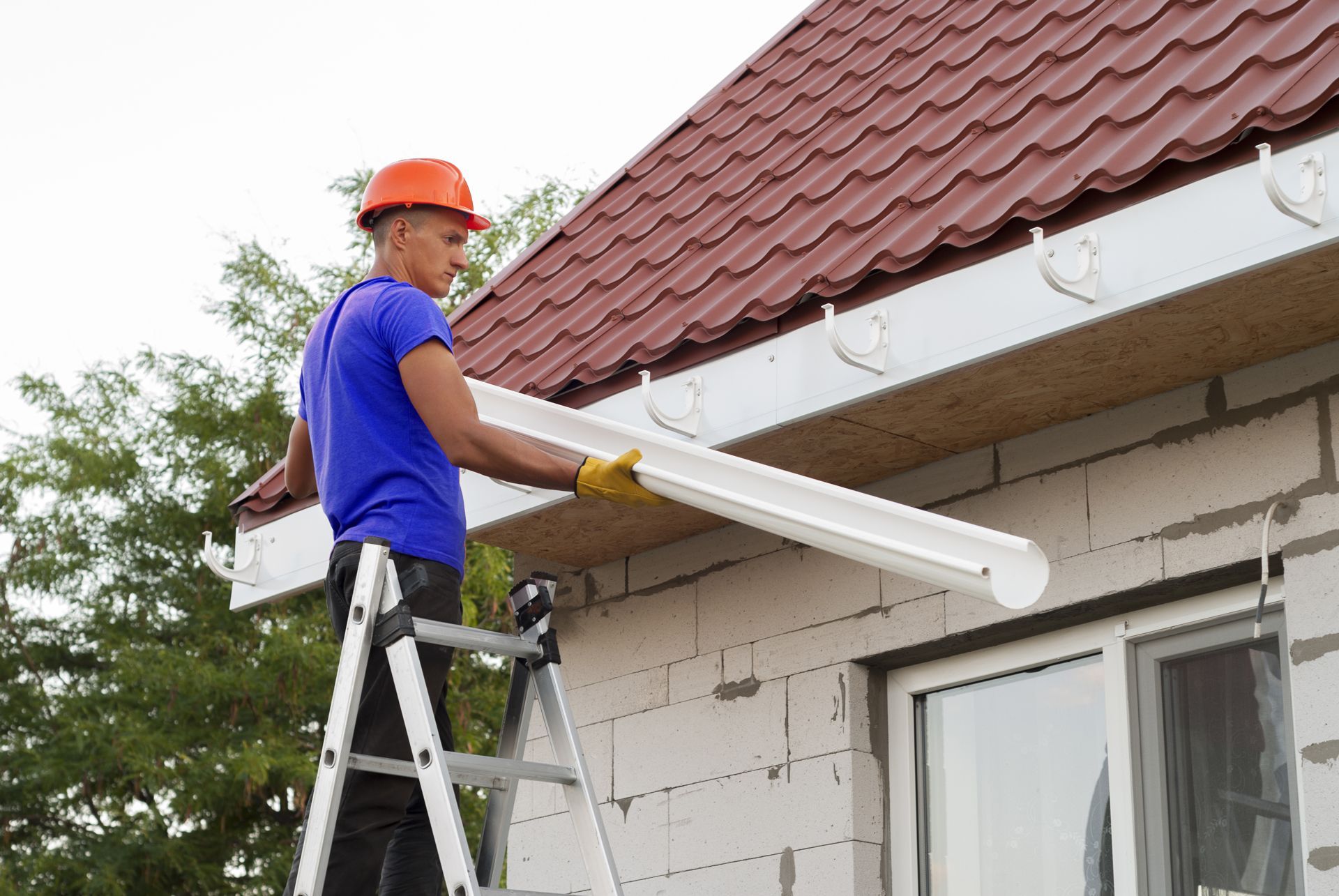 A man is standing on a ladder installing a gutter on the side of a house.