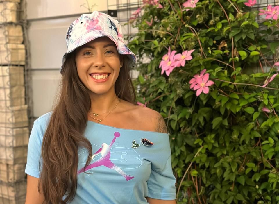 Woman with floral bucket hat and blue off-the-shoulder shirt smiles, near pink flowers and a brick wall.