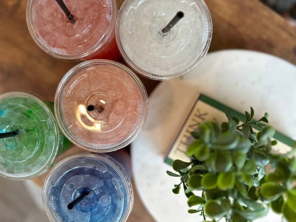 Colorful drinks in clear cups, black straws, on a wooden table with a plant and book.