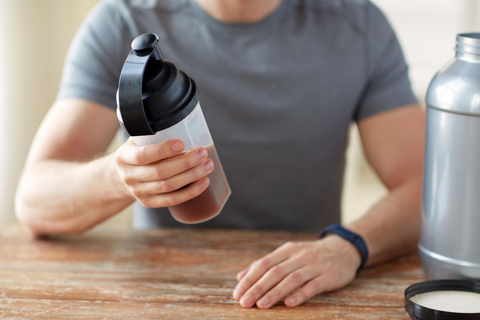 Man holding protein shake bottle, with large protein container and cup on a wooden table.