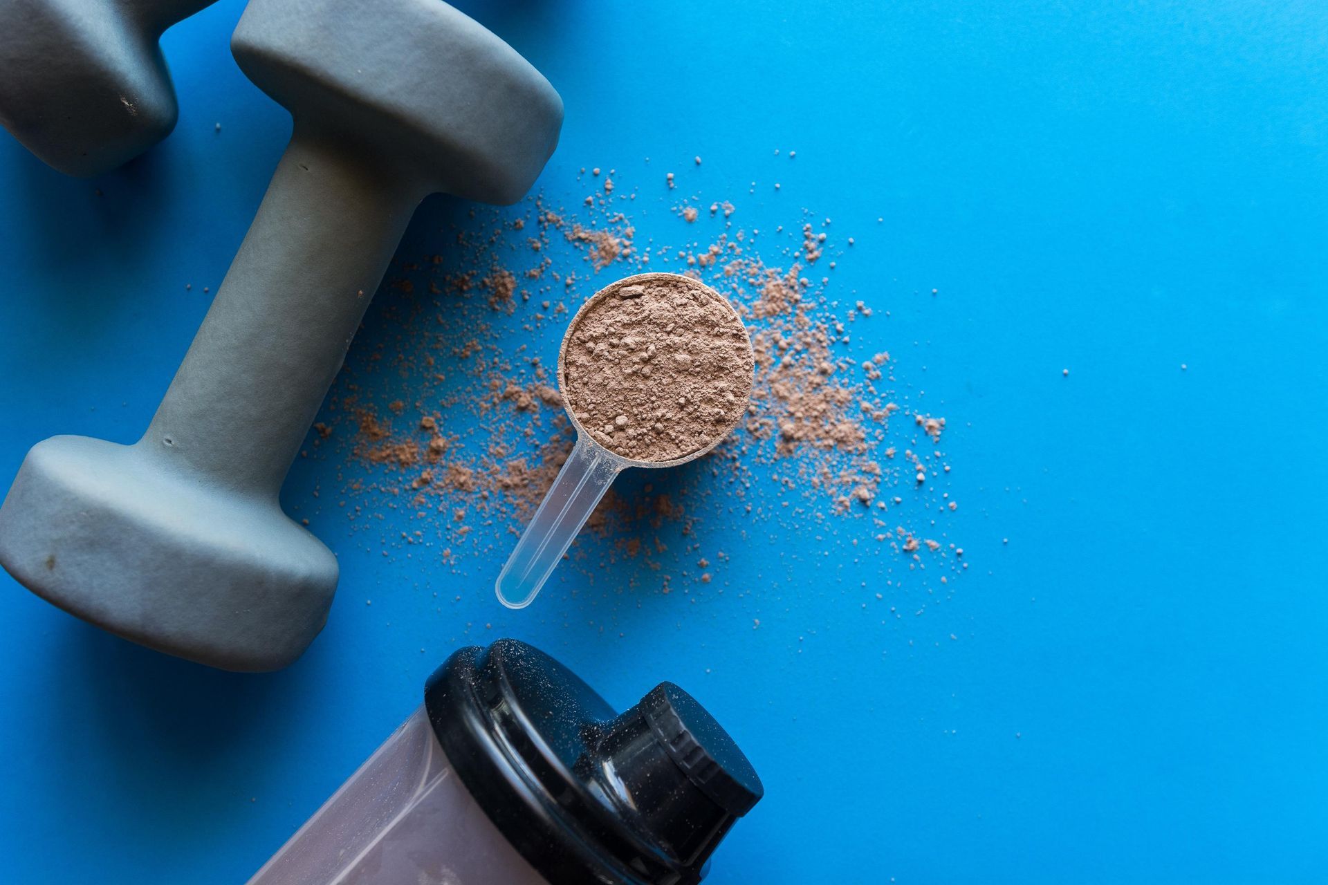 Dumbbells, protein powder scoop, and shaker bottle on a blue surface, suggesting fitness and nutrition.