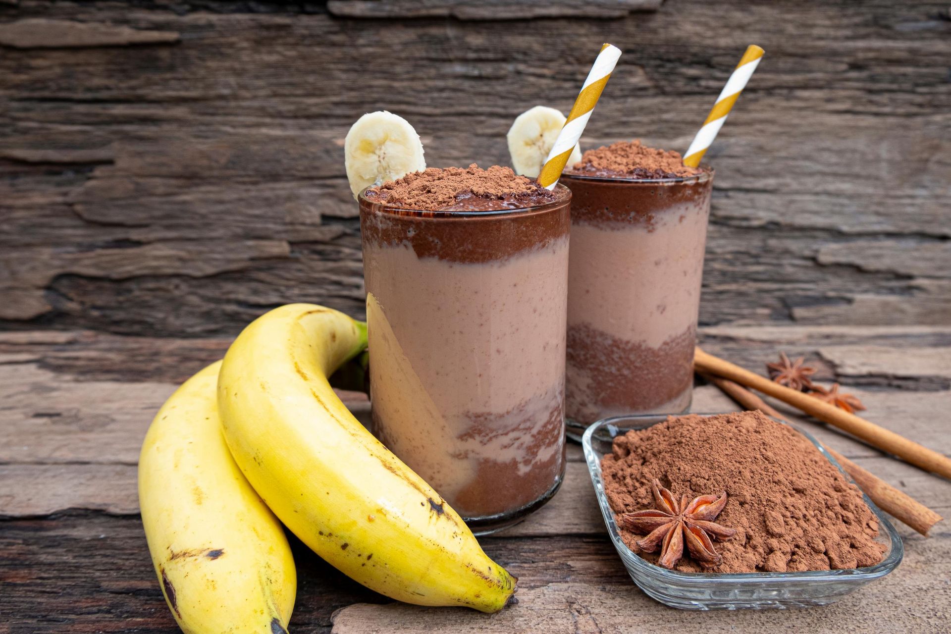 Two chocolate banana smoothies with bananas, cocoa, and straws on a wooden surface.