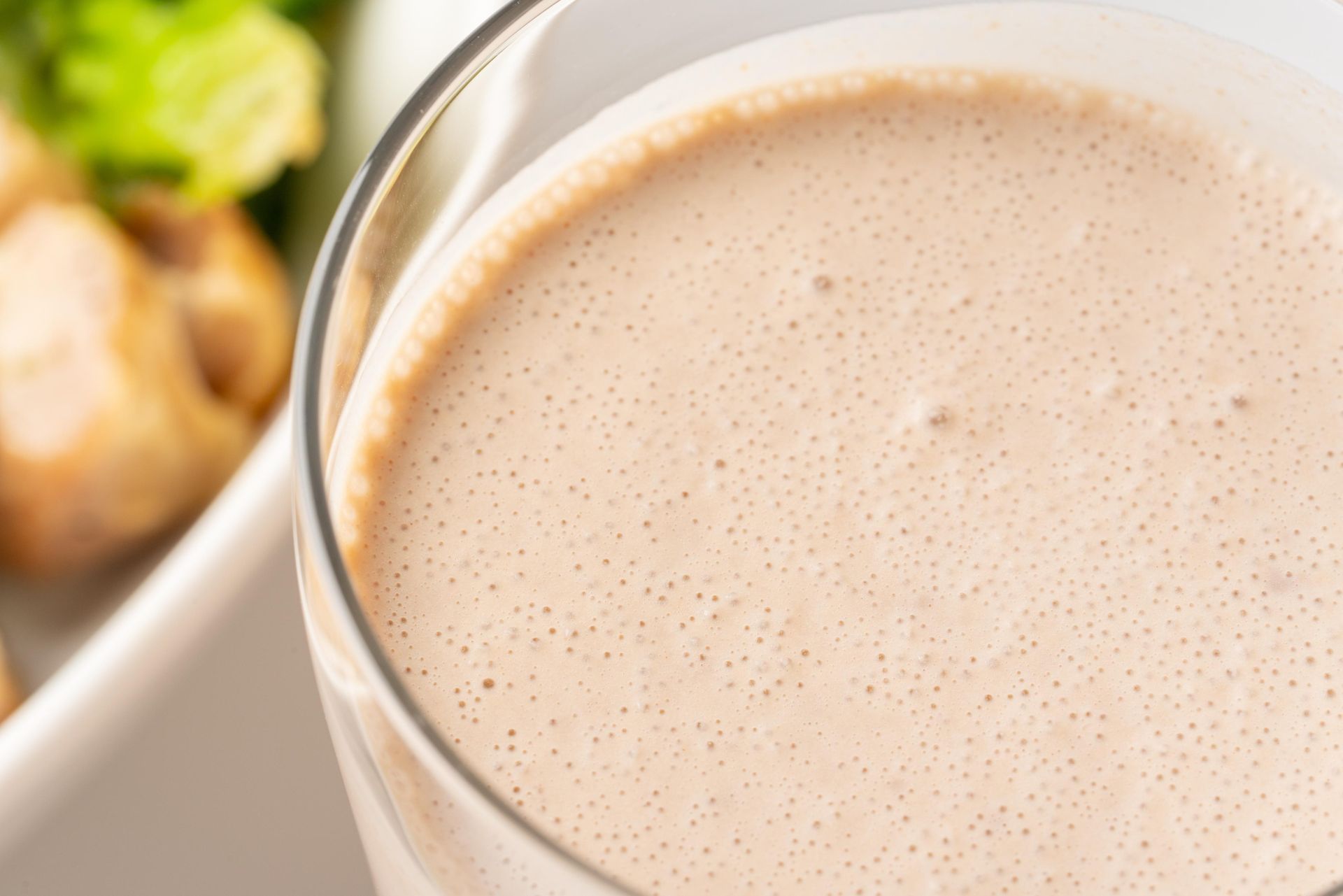 Frothy, light brown smoothie in a glass. Food items (broccoli, chicken) in the background.
