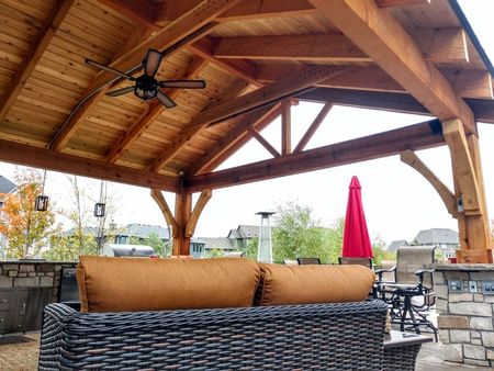 Covered outdoor patio with wicker sofa, stone fireplace, and red umbrella under a wooden roof