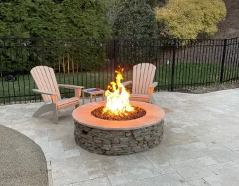 Outdoor stone fire pit with flames, surrounded by two Adirondack chairs on a patio.