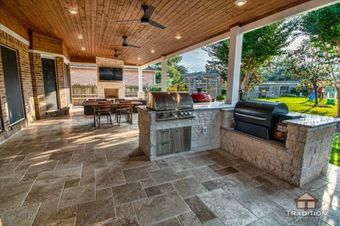Covered outdoor patio with stone tile floor, wood ceiling, dining table, grill, and view of a green yard