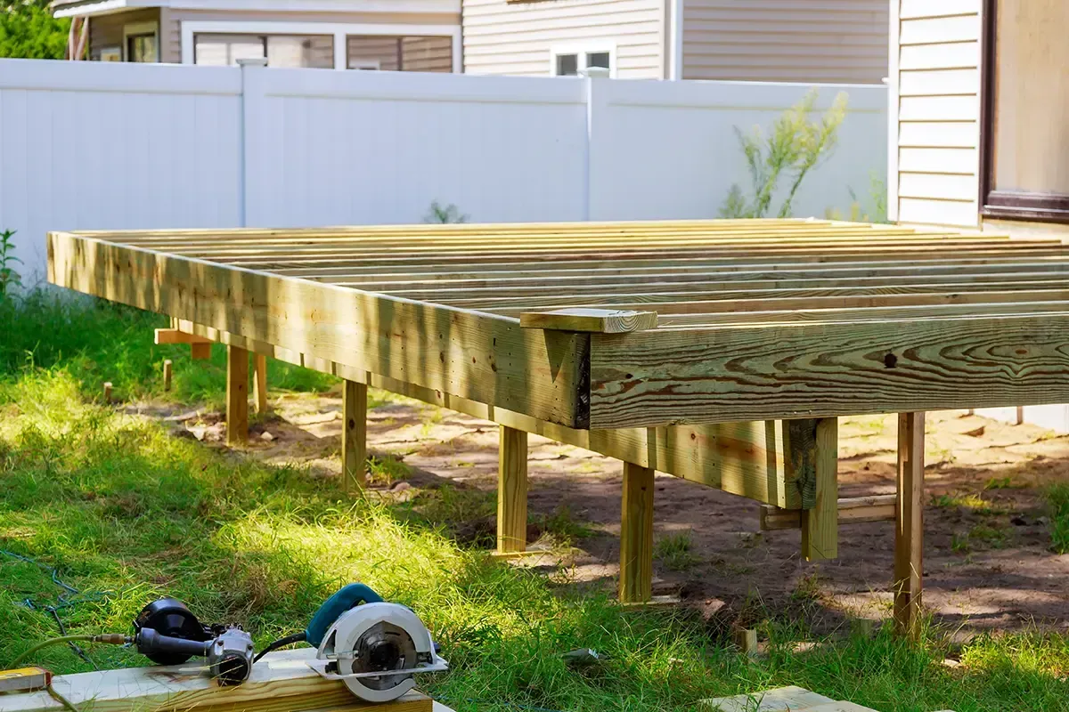Raised wooden deck in a backyard with grass and a fence, with tools on the ground nearby