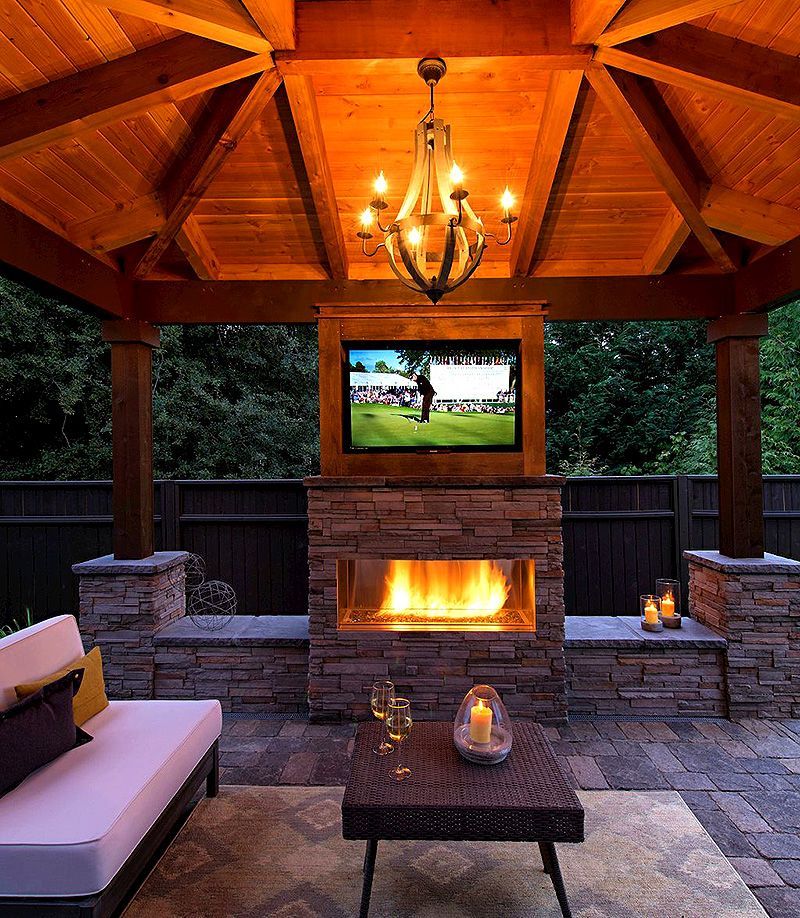 Cozy covered patio with stone fireplace, TV, chandelier, and cushioned seating at dusk