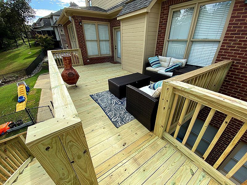 Small wooden patio deck with black outdoor seating, gray rug, and brick house exterior with windows.