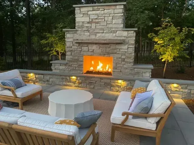 Outdoor stone fireplace with a lit fire, surrounded by cushioned patio seating and a round table at dusk