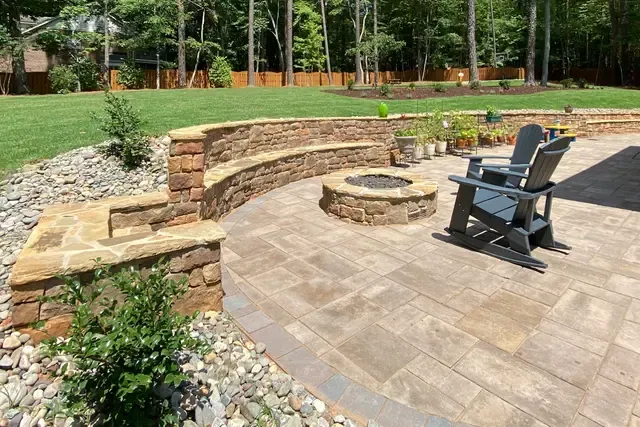 Stone patio with curved retaining wall, fire pit, and black chairs in a landscaped backyard