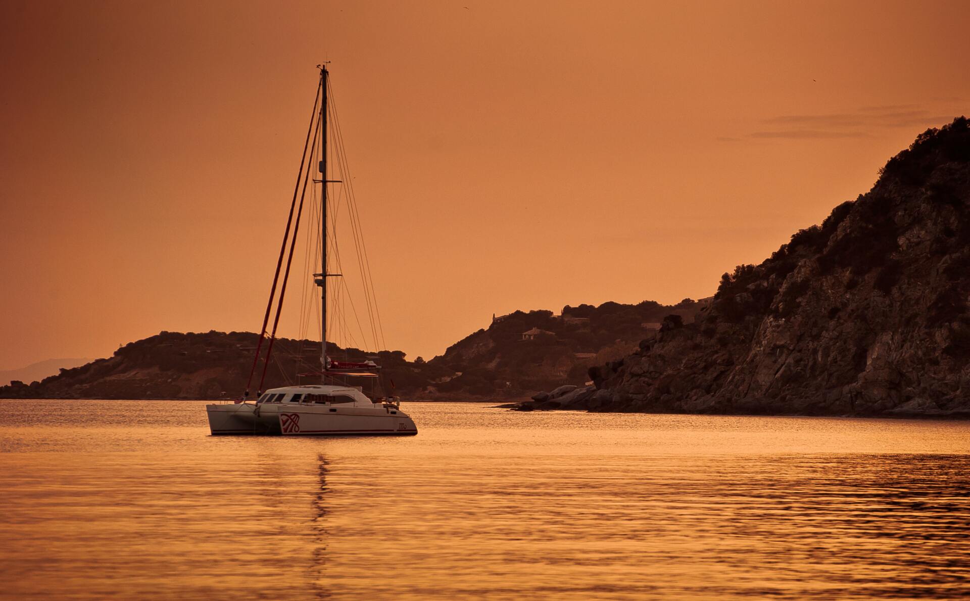 Sailboat anchored near a hilly coast during a warm, glowing sunset