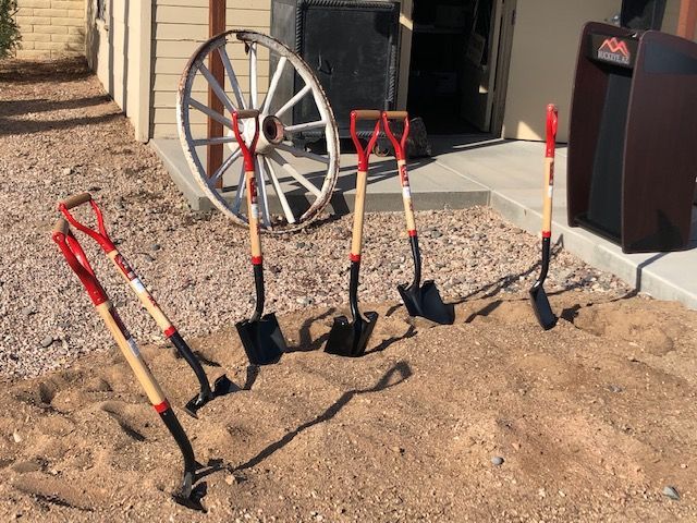 Four shovels are lined up in the sand in front of a wagon wheel.