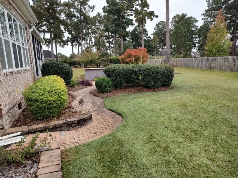 Curved brick walkway beside a house, trimmed shrubs, and a green lawn in a landscaped yard.
