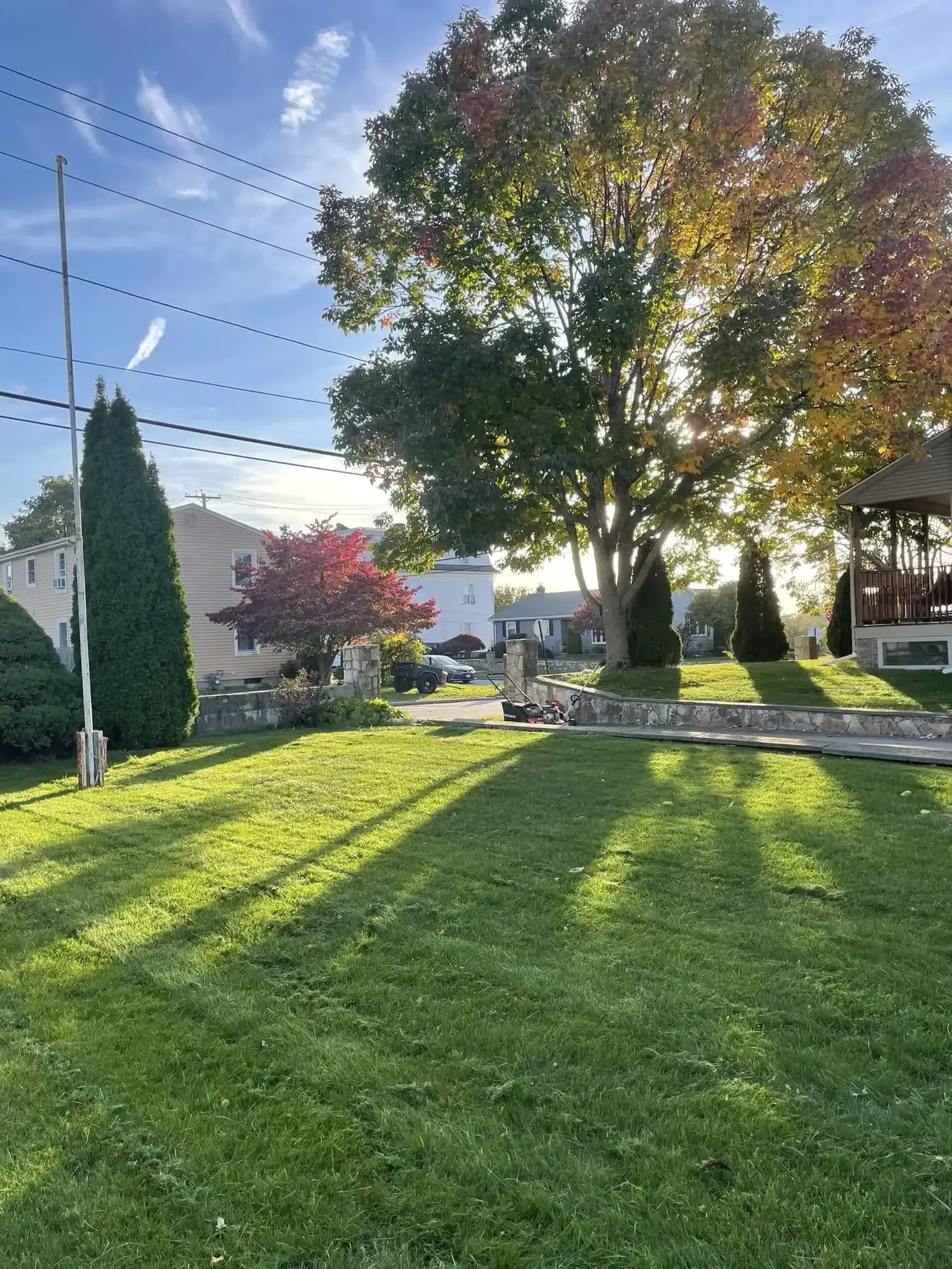 A grassy yard with a tall flagpole, trees in autumn colors, and a partially visible structure under a bright sunny sky.
