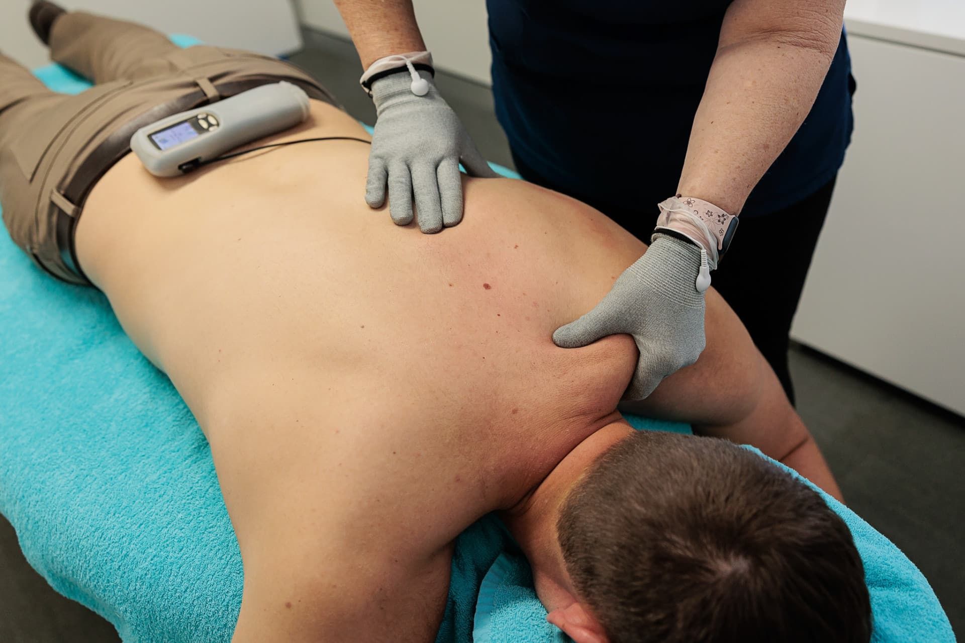 Man Receiving Back Massage with Electric Gloves, Lying on Turquoise Bed. Beige Pants, Dark Shirt — Grays Chiropractic & Wellness In Ooralea, QLD