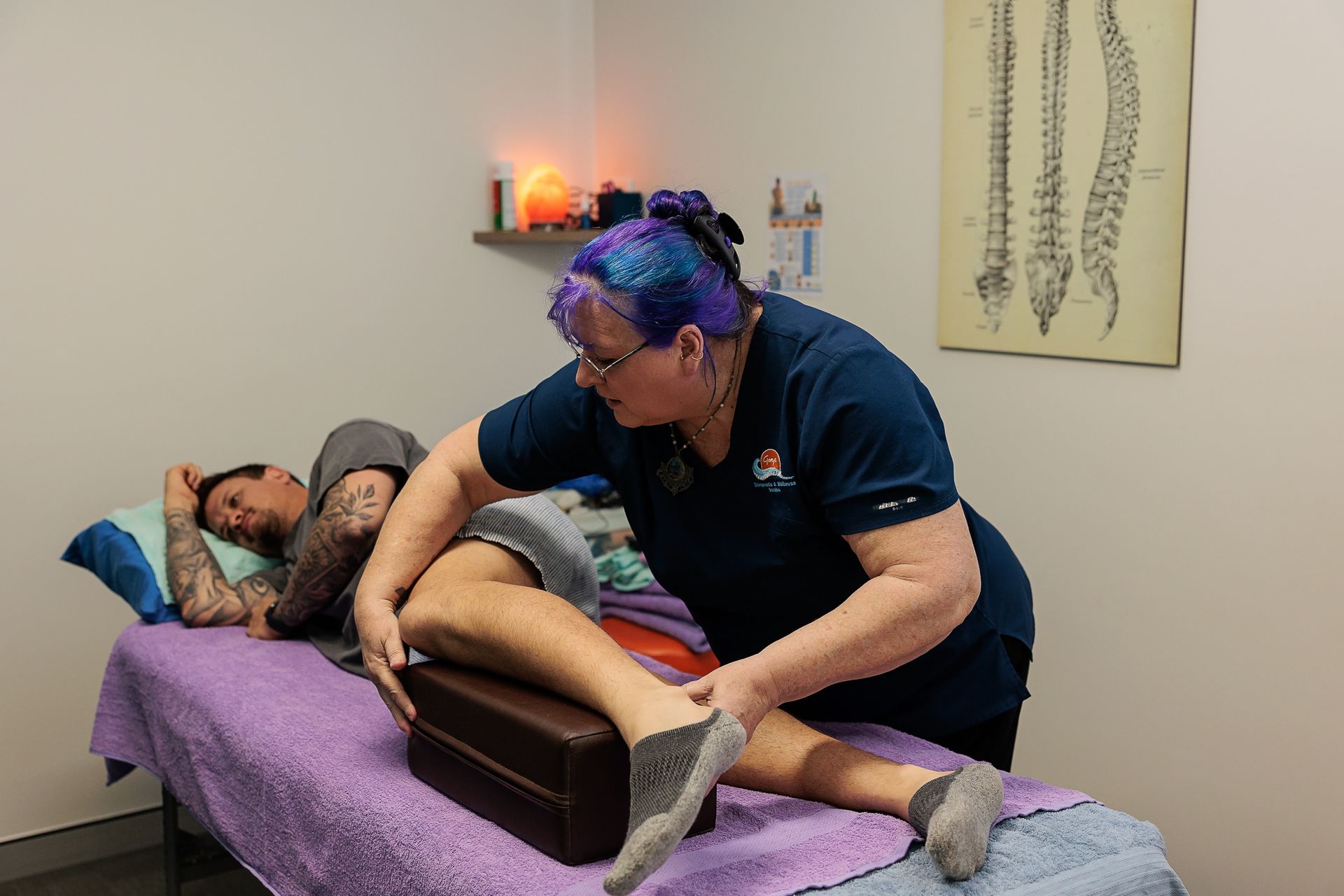 Woman in Blue Shirt Examines Man's Leg on Massage Table, Office Setting, Using a Block — Grays Chiropractic & Wellness In Paget, QLD