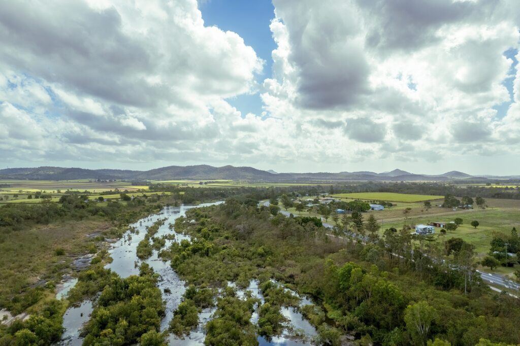 Aerial view of a meandering river with dense green vegetation under a cloudy sky and distant hills.— Grays Chiropractic & Wellness in Marian, QLD