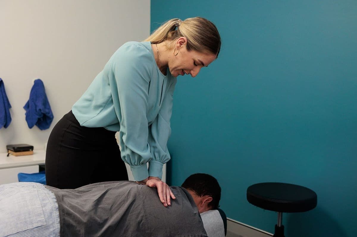 Woman in Blue Shirt Adjusting a Man's Back on An Examination Table. Teal Wall Backdrop — Grays Chiropractic & Wellness In Ooralea, QLD