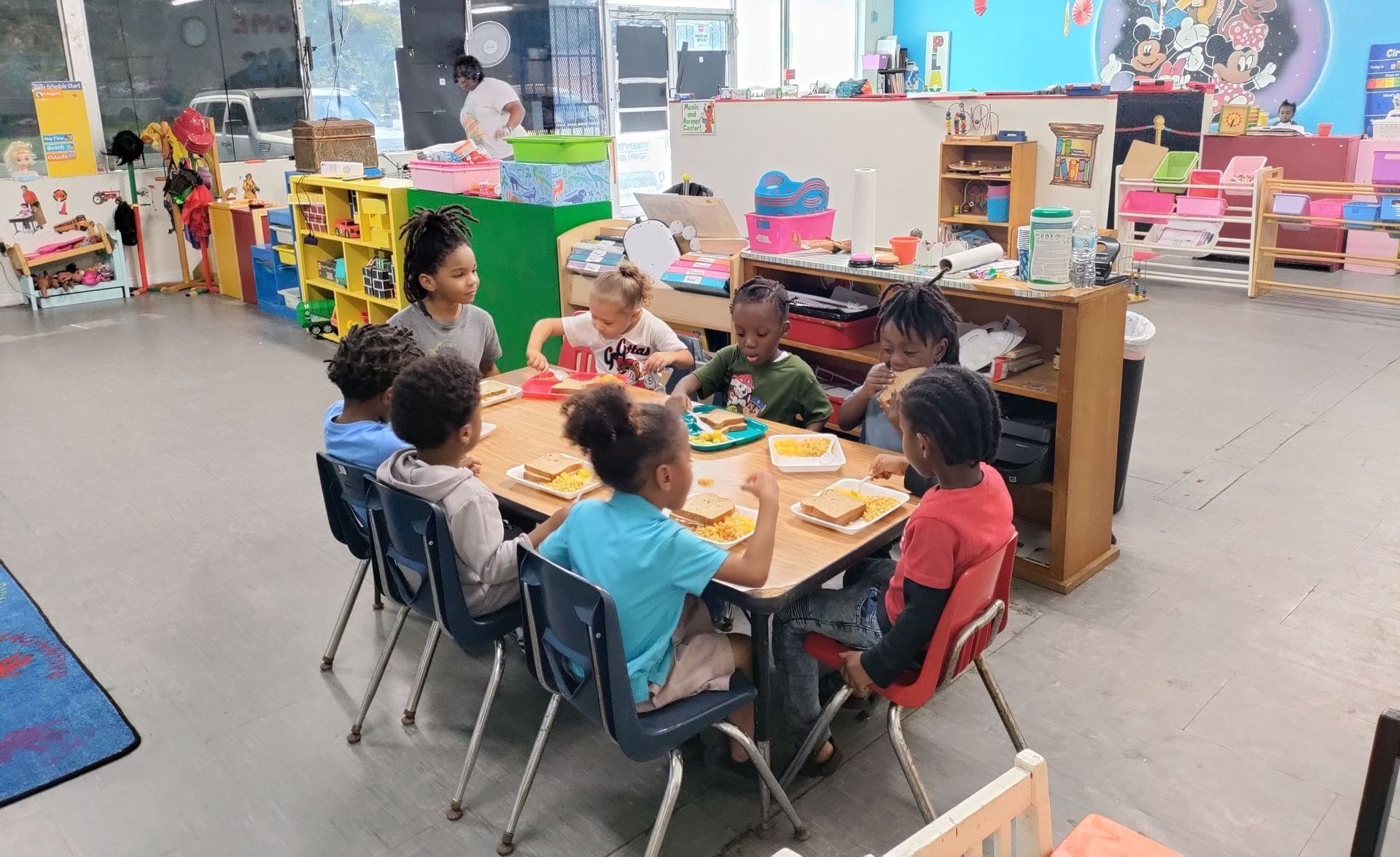 A group of children are sitting around a table in a classroom eating food.